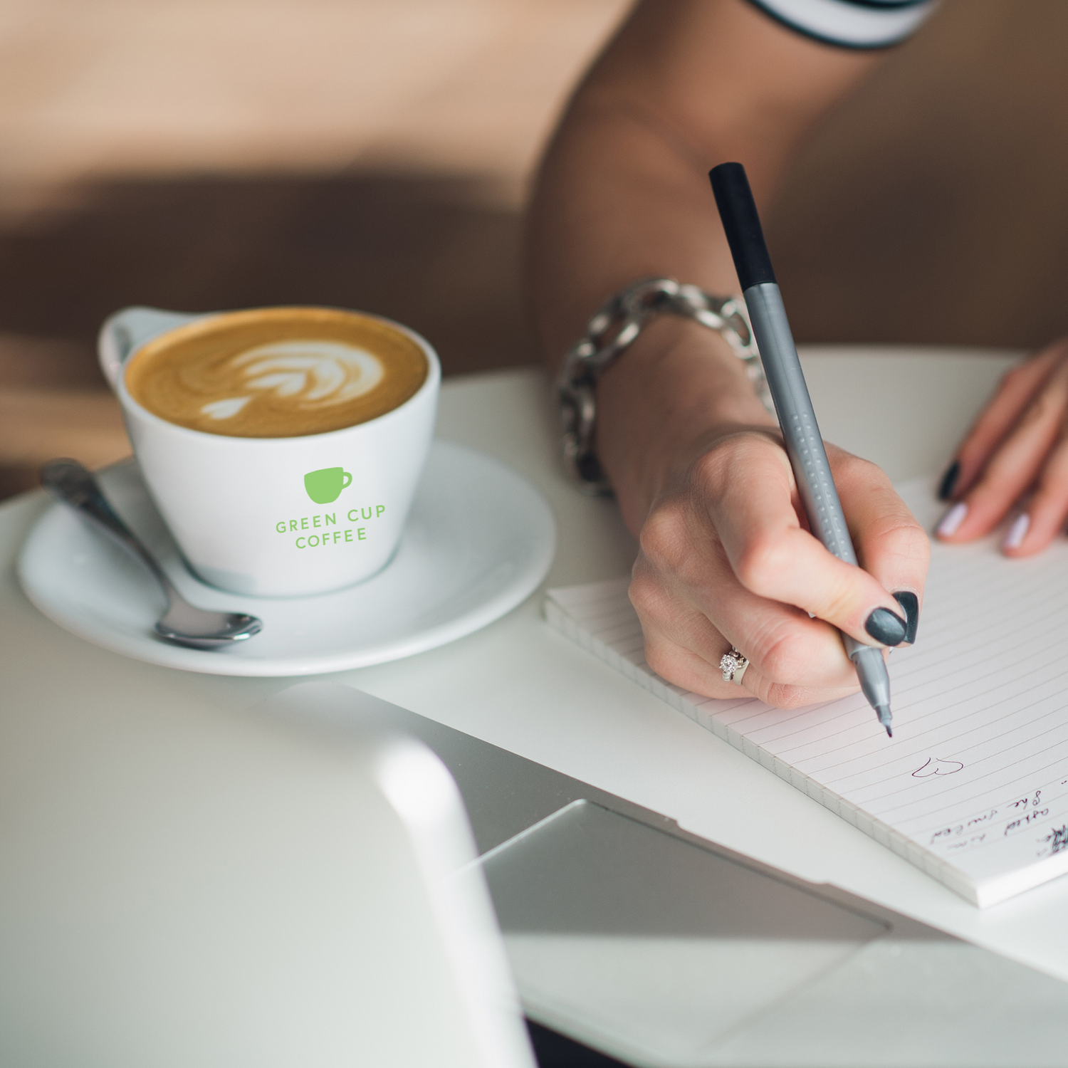 Hände einer Frau am Tisch im Büro mit Bürokaffee Cappuuccino mit Laptop, Block & Stift