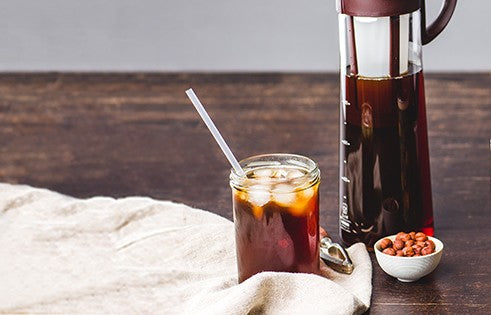 Ein Glas Cold Brew Kaffee mit Eiswürfeln, daneben eine Cold Brew Karaffe und eine Schale Kaffeebohnen auf einem Holztisch