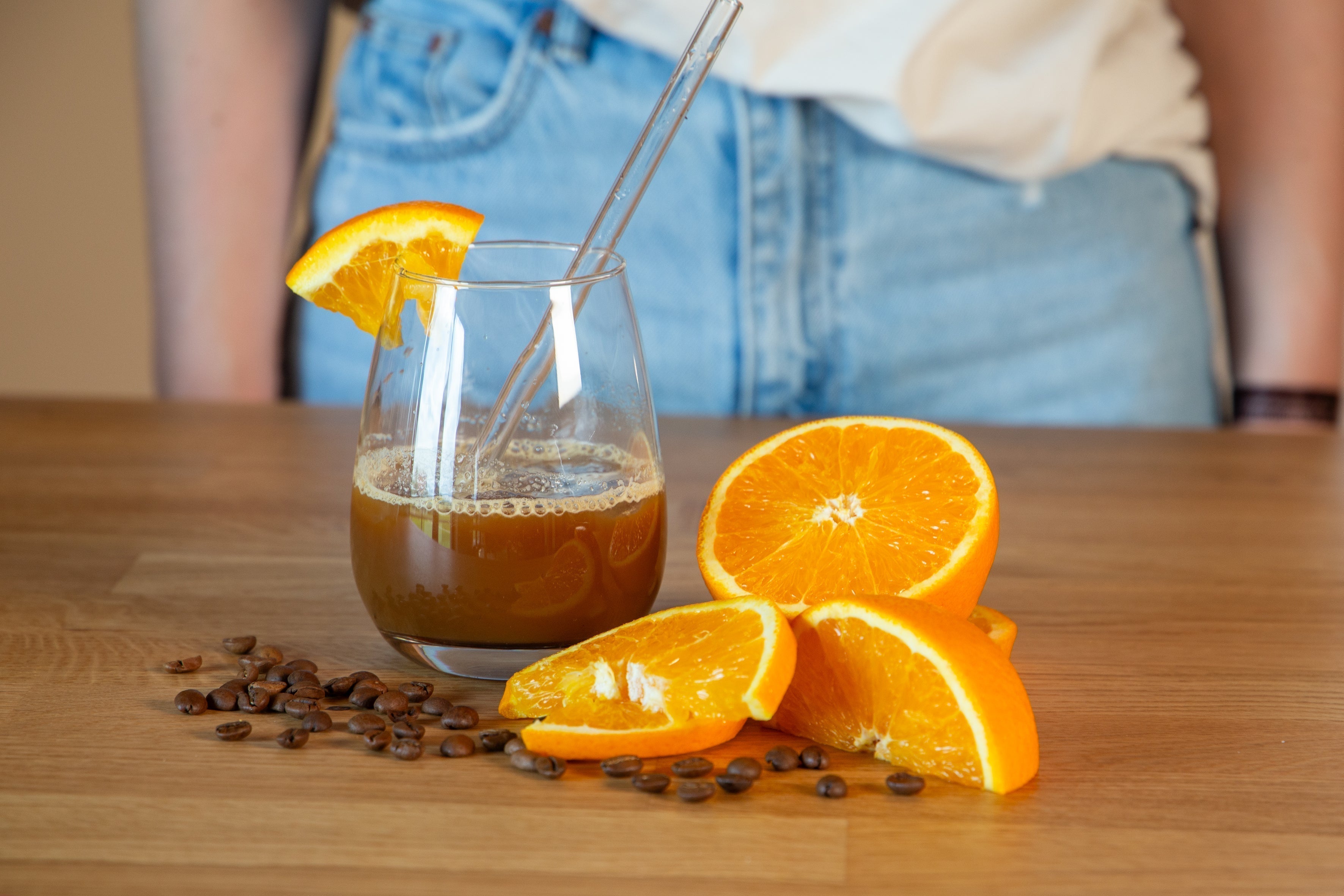 Iced Orange Espresso im Glas mit Orangenscheibe und Glastrinkhalm, dekoriert mit frischen Orangenspalten und Kaffeebohnen auf einem Holztisch – erfrischendes Sommergetränk mit fruchtiger Zitrusnote und Espresso