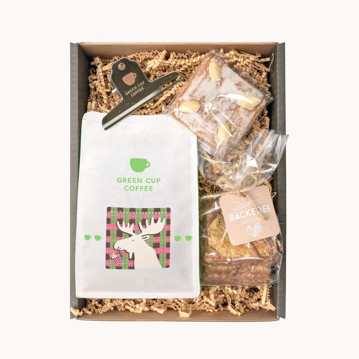 Geschenkbox mit einer Tüte Weihnachtsespresso, Mandelplättchen mit Schokoladenstreifen, einem Lebkuchen in Natur mit Mandeln und einem silbernen Kaffeetütenclip mit Green Cup Coffee Logo.