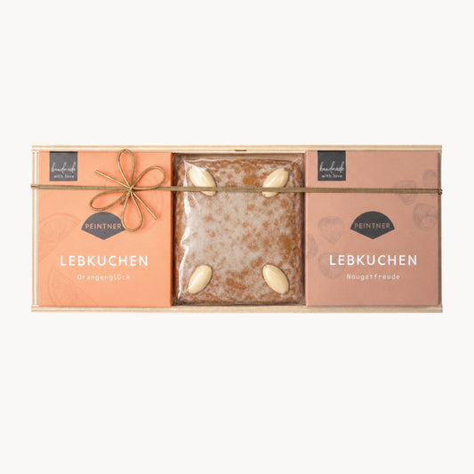 Lebkuchen-Set in einer Holzbox mit drei Teilen: links „Orangenglück“, in der Mitte ein Natur-Lebkuchen mit Mandeln, rechts „Nougatfreude“, alle Packungen mit Goldschleife.