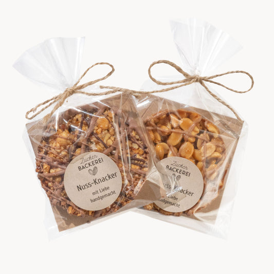 Zwei Tüten Nuss-Knacker mit Etikett „Zucker Bäckerei“, links mit gehackten Nüssen und Schokoladenstreifen, rechts mit ganzen Erdnüssen.